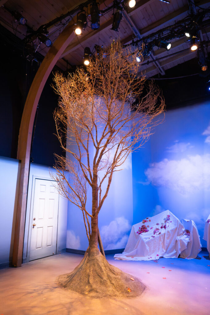 The Fall Show set: a bare tree rooted in a mound of earth, sky-painted walls, a white door and a sheet-draped couch