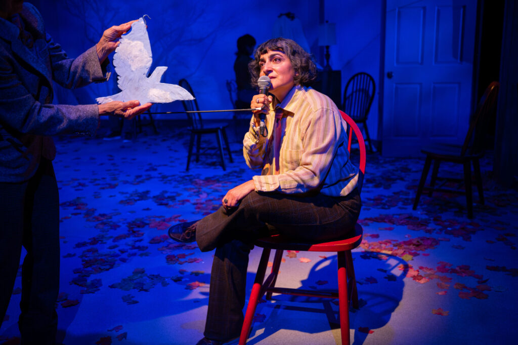 A performer on a red chair holds a microphone as a hand floats a white paper bird beside her