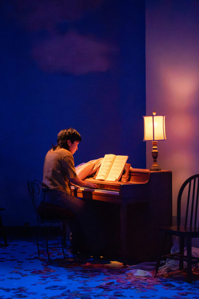 A musician plays an upright piano by lamplight under a painted sky