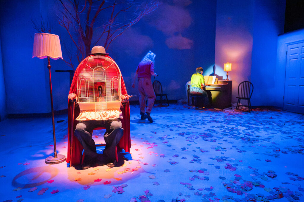 A seated performer in a bowler hat and red cloak, a lit birdcage where their chest should be