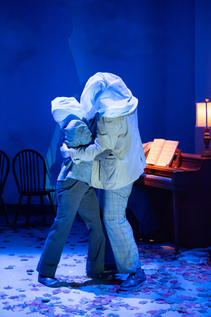 Two performers embrace beside a piano, their heads wrapped in white fabric