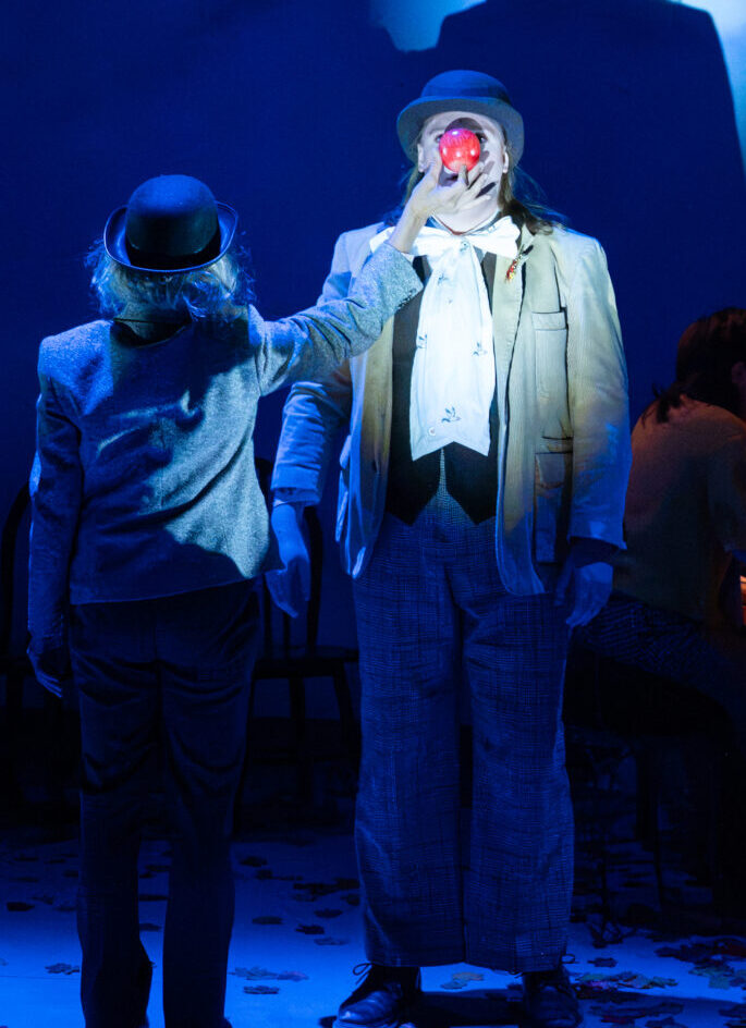 A performer in a bowler hat holds an apple in front of his face, his shadow cast large on the wall behind him